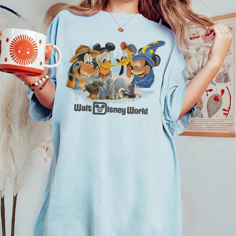 Pu&ograve; includere: T-shirt azzurra con una grafica di Topolino, Paperino, Pippo e un Topolino mago davanti a un castello, con la scritta "Walt Disney World". Una tazza bianca e arancione con un disegno di sole &egrave; tenuta in mano.
