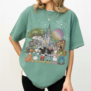 K&ouml;nnte beinhalten: Gr&uuml;nes T-Shirt mit einem farbenfrohen Grafikdesign eines Disney-Themenparks, einschlie&szlig;lich Schloss, Zug und Figuren wie Mickey und Minnie Mouse. Das Design zeigt auch Feuerwerk und eine gro&szlig;e Kugel.