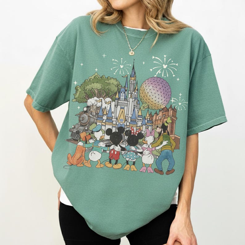 K&ouml;nnte beinhalten: Gr&uuml;nes T-Shirt mit einem farbenfrohen Grafikdesign eines Disney-Themenparks, einschlie&szlig;lich Schloss, Zug und Figuren wie Mickey und Minnie Mouse. Das Design zeigt auch Feuerwerk und eine gro&szlig;e Kugel.
