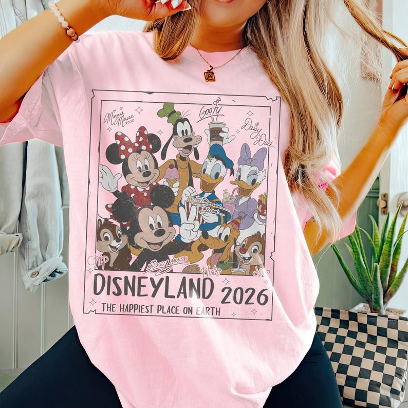 May include: Pink t-shirt featuring a Disneyland 2026 graphic with Mickey Mouse, Minnie Mouse, Goofy, Donald Duck, Daisy Duck, and Chip and Dale. The text "The Happiest Place on Earth" is also included.