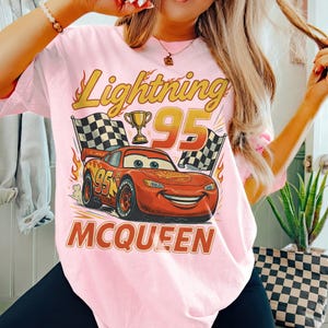 May include: Pale pink t-shirt with a graphic of Lightning McQueen from the film Cars. The design includes the text "Lightning 95", a trophy, checkered flags, and the name "McQueen".