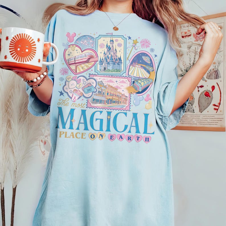 May include: Light blue t-shirt with a colorful graphic featuring various theme park attractions and the text "The most MAGICAL PLACE ON EARTH." A mug with a smiling sun design is held in the image.