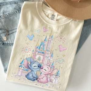 Könnte beinhalten: Cremefarbenes T-Shirt mit einer farbenfrohen Illustration eines Schlosses mit zwei Zeichentrickfiguren. Das Design umfasst Feuerwerk, Mickey Mouse-Ballons und Sterne. Das T-Shirt ist auf einer Jeans gefaltet.