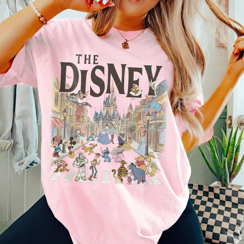 May include: A light pink t-shirt with a graphic of Disney characters crossing a street, with a castle in the background. The text "THE DISNEY" is printed above the graphic in a dark brown font.