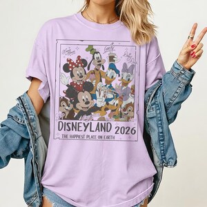 May include: A light purple t-shirt featuring a graphic of Disney characters including Mickey Mouse, Minnie Mouse, Donald Duck, and Goofy. The graphic reads "Disneyland 2026" and "The Happiest Place on Earth."