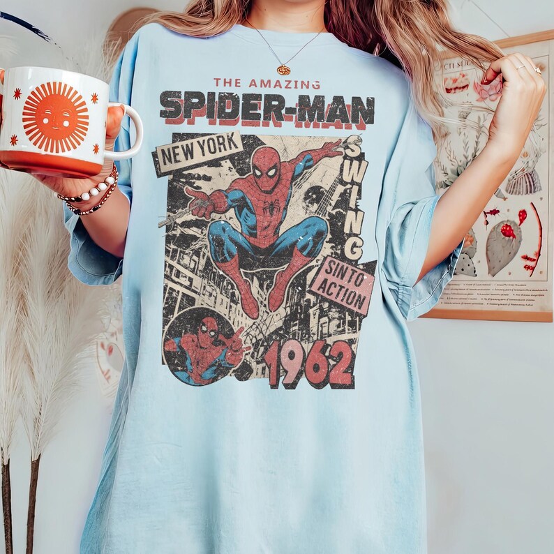 Op de afbeelding: Lichtblauw T-shirt met een vintage Spider-Man-ontwerp. Het ontwerp bevat de woorden "THE AMAZING SPIDER-MAN" en "NEW YORK", met een stripboekachtige illustratie van Spider-Man. Het jaar "1962" is ook zichtbaar.