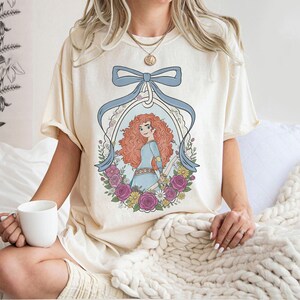 K&ouml;nnte beinhalten: Cremefarbenes T-Shirt mit einer Cartoon-Illustration einer Figur mit roten Locken, die ein blaues Kleid tr&auml;gt und von Blumen umgeben ist. Ein hellblaues Band umrahmt das Bild. Das T-Shirt wird von einer Person getragen.