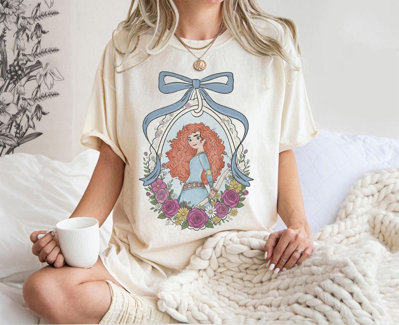 K&ouml;nnte beinhalten: Cremefarbenes T-Shirt mit einer Cartoon-Illustration einer Figur mit roten Locken, die ein blaues Kleid tr&auml;gt und von Blumen umgeben ist. Ein hellblaues Band umrahmt das Bild. Das T-Shirt wird von einer Person getragen.