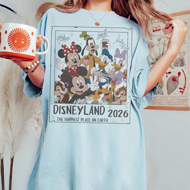May include: Light blue t-shirt featuring a Disneyland 2026 graphic with Mickey Mouse, Minnie Mouse, Goofy, Donald Duck, Daisy Duck, Chip, and Dale. The text "The Happiest Place on Earth" is also included.