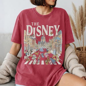 May include: Red t-shirt with the text "THE DISNEY" and a colourful illustration of Disney characters crossing a street. The design includes Mickey Mouse, Cinderella, Buzz Lightyear, and other characters.