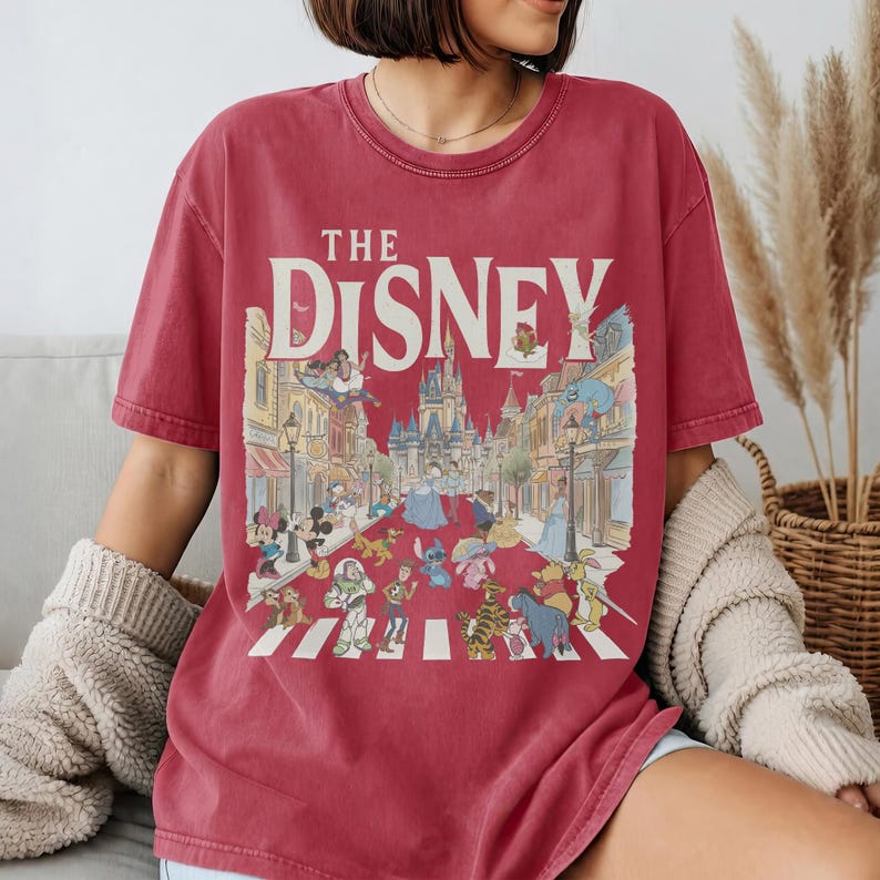 May include: Red t-shirt with the text "THE DISNEY" and a colourful illustration of Disney characters crossing a street. The design includes Mickey Mouse, Cinderella, Buzz Lightyear, and other characters.