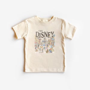 May include: A cream-coloured t-shirt with a colourful illustration of a Disney theme park scene and cartoon characters. The text "THE DISNEY" is printed above the illustration in dark brown. The shirt has short sleeves and a crew neck.