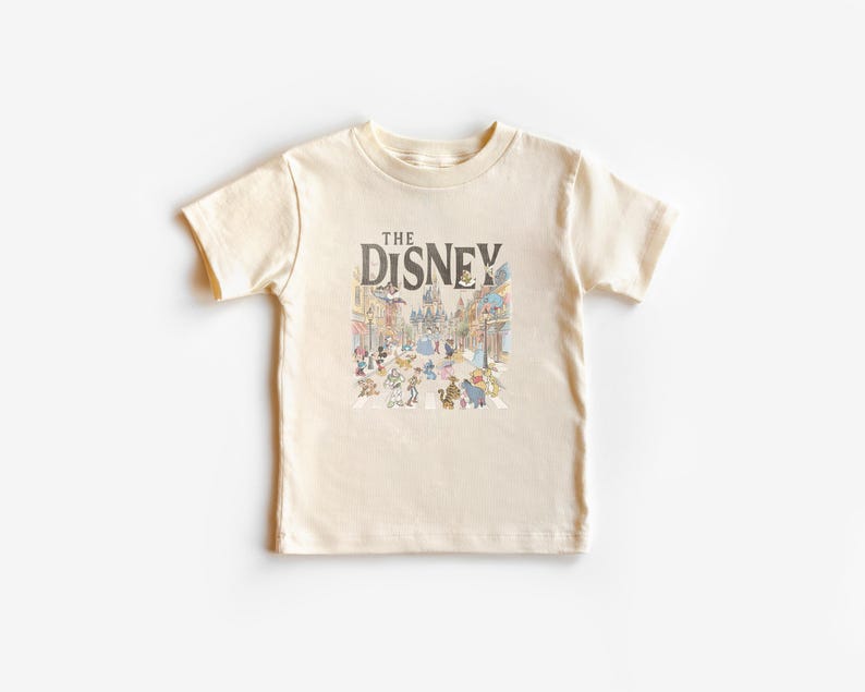 May include: A cream-coloured t-shirt with a colourful illustration of a Disney theme park scene and cartoon characters. The text "THE DISNEY" is printed above the illustration in dark brown. The shirt has short sleeves and a crew neck.