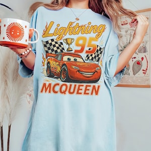 May include: Pale blue t-shirt with a graphic of Lightning McQueen from the Cars film. The design includes the text "Lightning 95" and "McQueen", with checkered flags and a trophy. A mug with a sun design is held in the image.