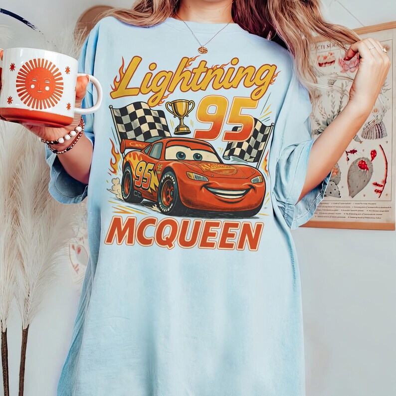 May include: Pale blue t-shirt with a graphic of Lightning McQueen from the Cars film. The design includes the text "Lightning 95" and "McQueen", with checkered flags and a trophy. A mug with a sun design is held in the image.