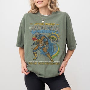 May include: Olive green t-shirt featuring a graphic of The Mandalorian from Star Wars. The design includes the Mandalorian character, Grogu, and the text "STAR WARS THE MANDALORIAN". The shirt also has the text "WHEN YOU CHOOSE TO WALK THE WAY OF THE MANDALONE YOU ARE BOTH HUNTER & PREY."