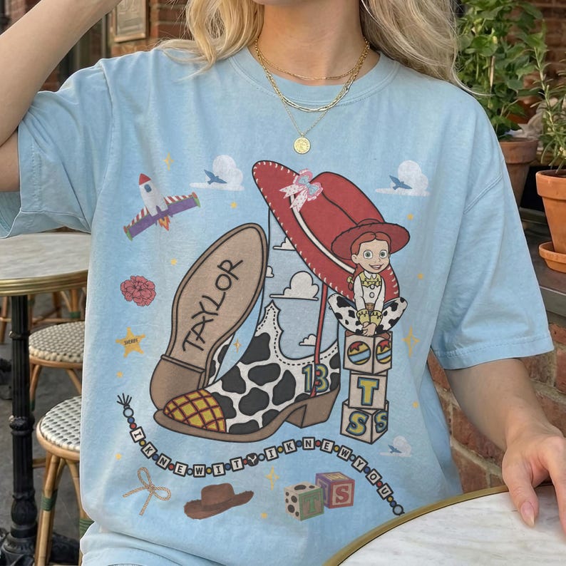 May include: A light blue t-shirt featuring a graphic print of a cow-print cowboy boot with the name Taylor on the sole, a red hat, a character, and blocks with the letters T, S, and 13. The design includes a rocket, stars, and beads with text.