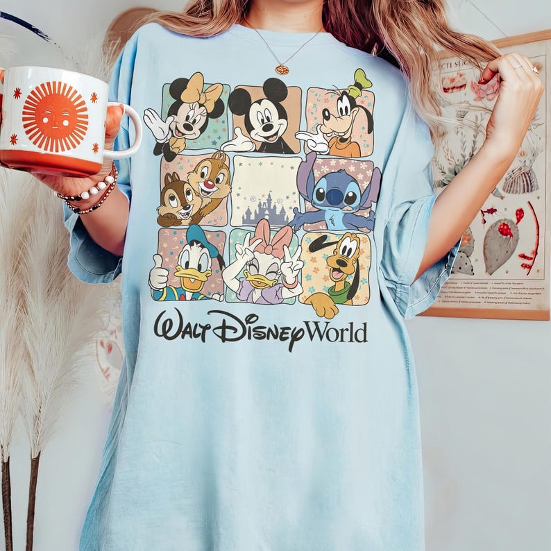 K&ouml;nnte beinhalten: Hellblaues T-Shirt mit einem Rastermuster, das Disney-Figuren wie Mickey und Minnie Mouse, Donald Duck und Stitch zeigt. Der Schriftzug "Walt Disney World" ist in schwarzer Schrift gedruckt.