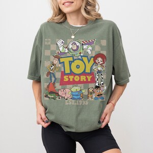 May include: Green short-sleeve t-shirt featuring a graphic of Toy Story characters, including Buzz Lightyear, Woody, and Jessie. The word "TOY STORY" is in yellow and red, with "EST.1995" below. The shirt has a vintage, worn look.