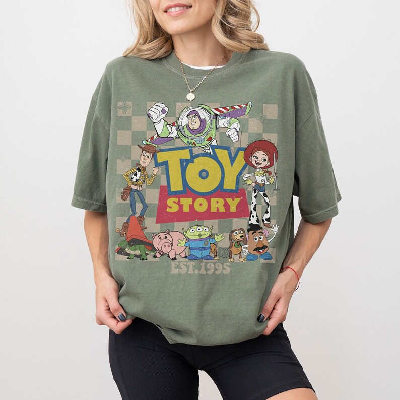 May include: Green short-sleeve t-shirt featuring a graphic of Toy Story characters, including Buzz Lightyear, Woody, and Jessie. The word "TOY STORY" is in yellow and red, with "EST.1995" below. The shirt has a vintage, worn look.