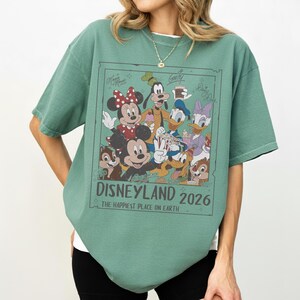 May include: Green t-shirt featuring a graphic of Disney characters including Mickey Mouse, Minnie Mouse, Goofy, Donald Duck, and Daisy Duck. The graphic is framed with the text "DISNEYLAND 2026" and "THE HAPPIEST PLACE ON EARTH."