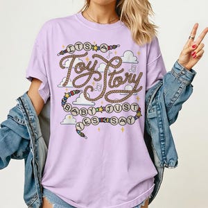 May include: A light purple t-shirt featuring a graphic print with the text "ITS A Toy Story BABY JUST YES SAY". The design includes rope-style lettering, colorful beads, yellow stars, musical notes, and white clouds on a lavender fabric background.