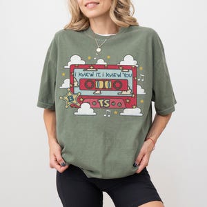 May include: An olive green t-shirt featuring a graphic of a red cassette tape with the text "I KNEW IT, I KNEW YOU" and "TS". The design includes white clouds, yellow stars, and musical notes surrounding the cassette tape on the front.