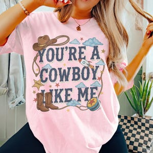 Puede incluir: Una camiseta rosa claro con un estampado gr&aacute;fico que incluye el texto "YOU'RE A COWBOY LIKE ME" en letras azul oscuro. El dise&ntilde;o presenta un sombrero de vaquero marr&oacute;n, una estrella de sheriff, botas vaqueras marrones, un cohete y un lazo.