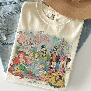 May include: Cream-colored t-shirt featuring a colorful graphic of Disney characters, including Dumbo, Mickey Mouse, and Ariel. The text "Magic Kingdom Walt Disney World" is printed below the characters. The shirt is folded on top of blue jeans.