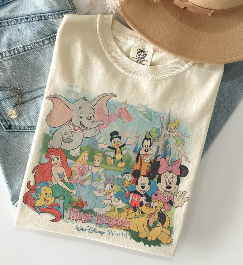 May include: Cream-colored t-shirt featuring a colorful graphic of Disney characters, including Dumbo, Mickey Mouse, and Ariel. The text "Magic Kingdom Walt Disney World" is printed below the characters. The shirt is folded on top of blue jeans.