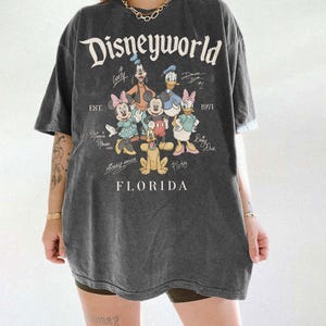 K&ouml;nnte beinhalten: Dunkelgraues T-Shirt mit dem Schriftzug "Disneyworld" und einer Grafik von Disney-Figuren, darunter Mickey Mouse, Minnie Mouse, Goofy, Donald Duck und Pluto. Das Shirt hat auch den Text "EST. 1971" und "Florida".