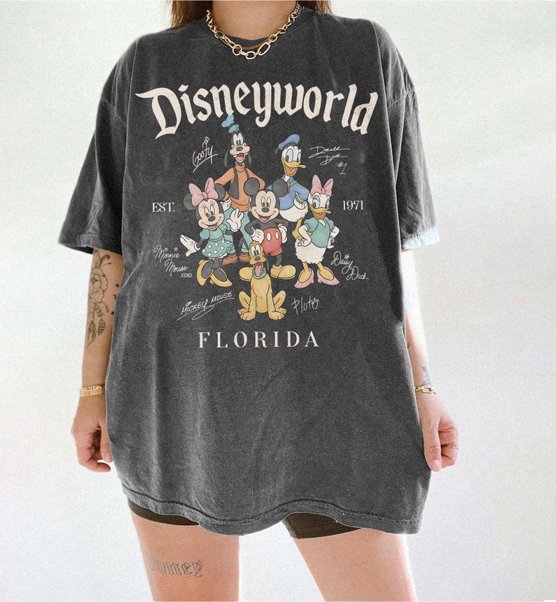 K&ouml;nnte beinhalten: Dunkelgraues T-Shirt mit dem Schriftzug "Disneyworld" und einer Grafik von Disney-Figuren, darunter Mickey Mouse, Minnie Mouse, Goofy, Donald Duck und Pluto. Das Shirt hat auch den Text "EST. 1971" und "Florida".