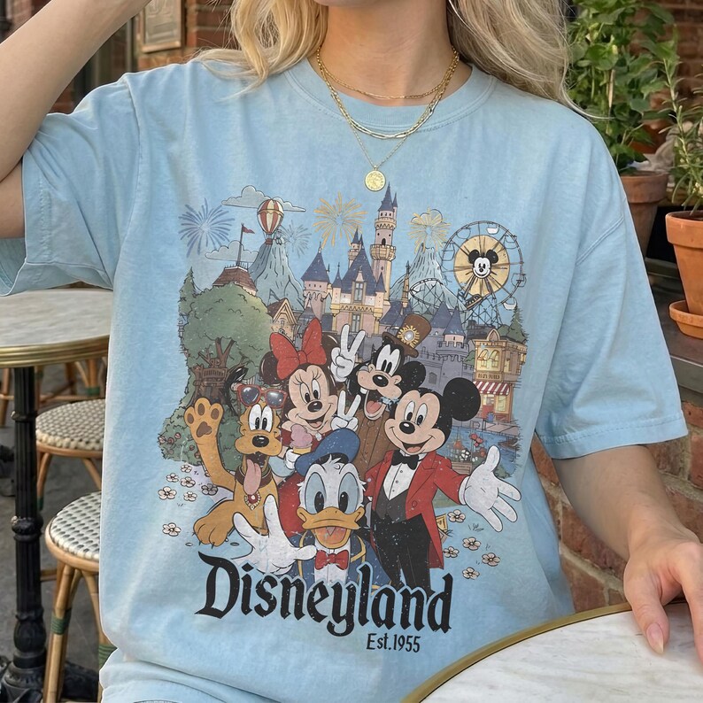May include: Light blue t-shirt featuring a Disneyland graphic with Mickey Mouse, Minnie Mouse, Donald Duck, Pluto, and Goofy. The graphic includes a castle, Ferris wheel, and the text "Disneyland Est.1955".