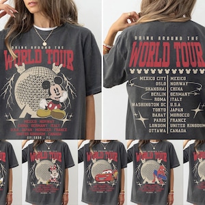 May include: Dark gray t-shirt with a graphic of Mickey Mouse in front of a geometric sphere, with the text "DRINK AROUND THE WORLD TOUR". The back of the shirt lists cities and countries. Additional shirts feature other Disney characters.