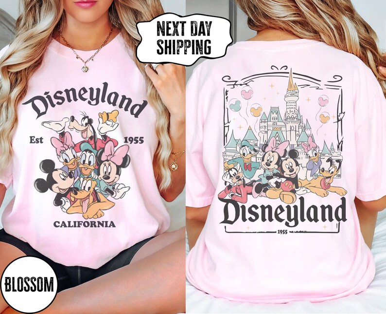May include: A light pink t-shirt featuring the Disneyland logo and cartoon characters on the front and back. The front design includes Mickey Mouse, Minnie Mouse, Donald Duck, Daisy Duck, and Goofy. The back design shows the Disneyland castle.