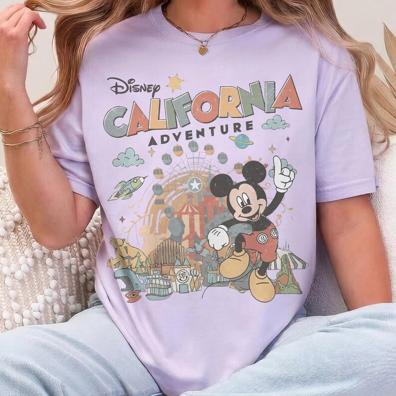 Puede incluir: Una camiseta de color lila claro con un gr&aacute;fico de Mickey Mouse y las palabras "Disney California Adventure". El dise&ntilde;o incluye una noria, un cohete y otros elementos de parque de atracciones. La camiseta es de estilo c&oacute;modo e informal.