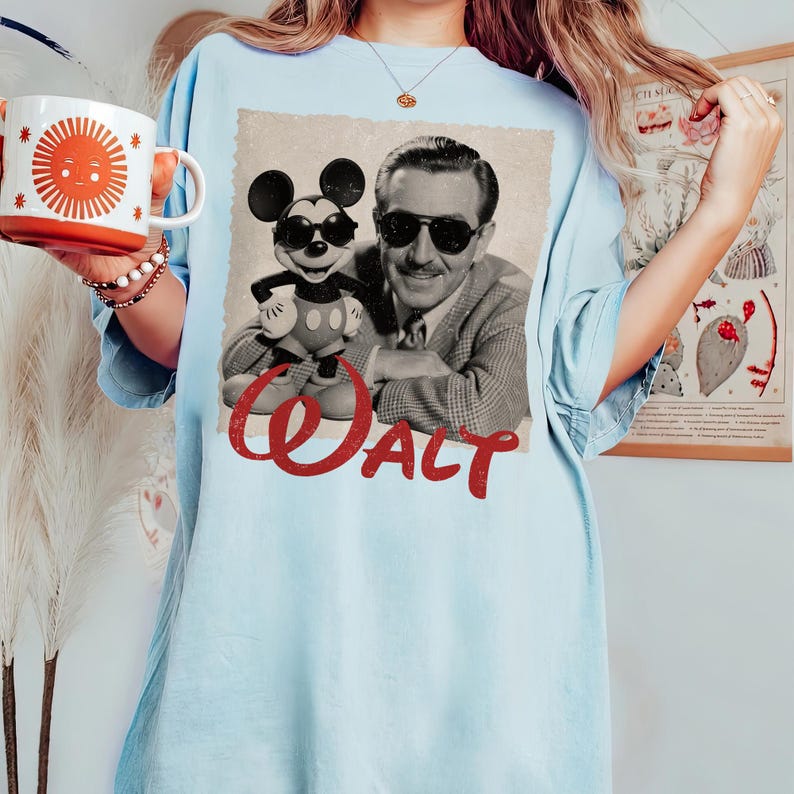 May include: Light blue t-shirt featuring a vintage-style image of Walt Disney with Mickey Mouse wearing sunglasses. The word "Walt" is printed in red script across the bottom. A mug with a sun design is held in the hand.