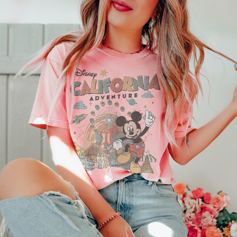 Puede incluir: Camiseta rosa claro con la inscripci&oacute;n "Disney California Adventure" y un dibujo de Mickey Mouse. El dise&ntilde;o incluye una noria, nubes y planetas. La camiseta tiene mangas cortas y cuello redondo.