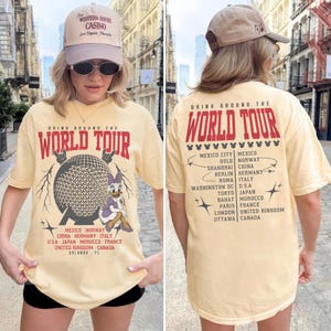 May include: A light yellow t-shirt with the text "DRINK AROUND THE WORLD TOUR" in red. The front features a graphic of a globe and Daisy Duck. The back lists cities. The person is wearing a beige cap.