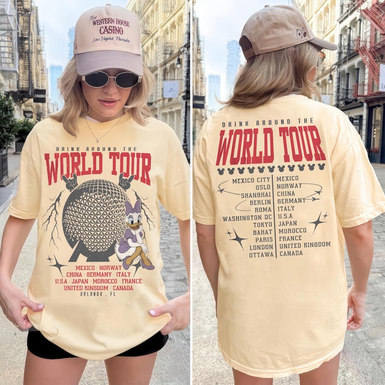 May include: A light yellow t-shirt with the text "DRINK AROUND THE WORLD TOUR" in red. The front features a graphic of a globe and Daisy Duck. The back lists cities. The person is wearing a beige cap.
