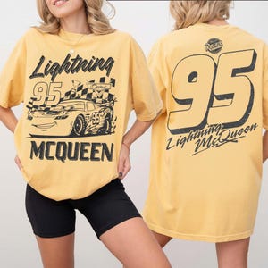 May include: Mustard yellow t-shirt featuring a graphic of Lightning McQueen from the Cars movie. The front of the shirt displays the text "Lightning McQueen" and "95" with a race car design. The back of the shirt has a large "95" and "Lightning McQueen" text.