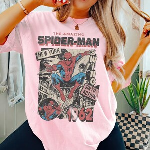 Op de afbeelding: Lichtroze t-shirt met een vintage Spider-Man-afbeelding. Het ontwerp bevat de woorden "THE AMAZING SPIDER-MAN", "NEW YORK" en "1962". De superheld wordt afgebeeld in een dynamische pose, met een retro stripboekstijl.