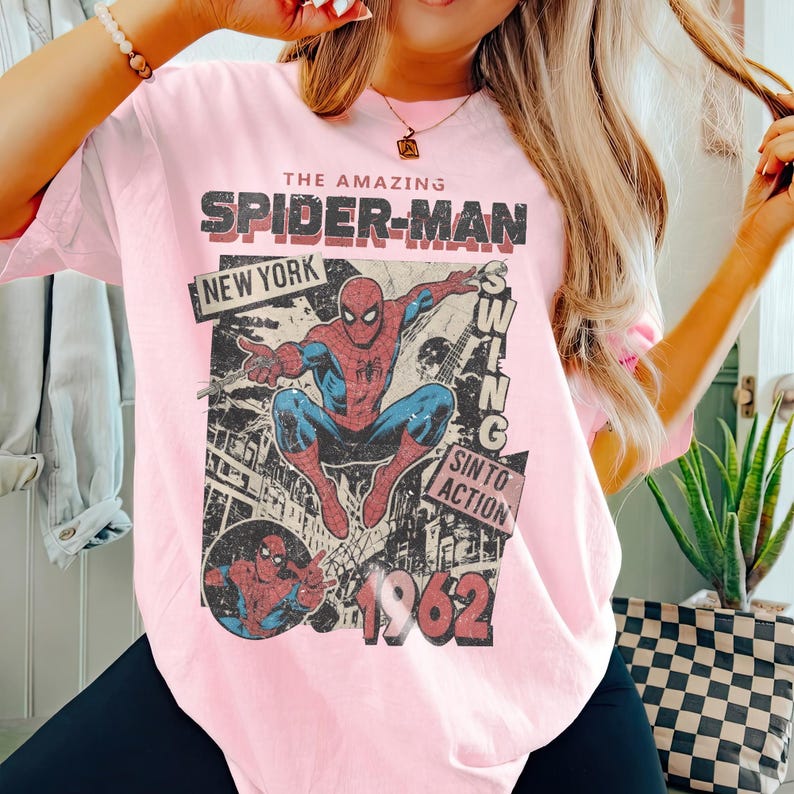 Op de afbeelding: Lichtroze t-shirt met een vintage Spider-Man-afbeelding. Het ontwerp bevat de woorden "THE AMAZING SPIDER-MAN", "NEW YORK" en "1962". De superheld wordt afgebeeld in een dynamische pose, met een retro stripboekstijl.