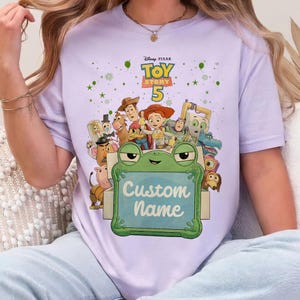 Puede incluir: Una camiseta lila con el logo de Disney Pixar Toy Story 5 y personajes. Un marco verde con forma de rana con las palabras "Custom Name" en primer plano. La camiseta tiene cuello redondo.