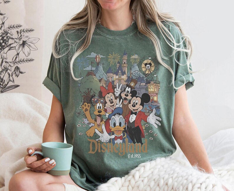 May include: Green t-shirt featuring a colorful graphic of Disney characters including Mickey Mouse, Minnie Mouse, and Goofy. The graphic depicts a park scene with fireworks and the text "Disneyland Est. 1955". The shirt is worn by a person.