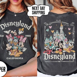 May include: Dark gray t-shirt featuring the Disneyland logo with Mickey Mouse, Minnie Mouse, Donald Duck, Daisy Duck, and Goofy. The front of the shirt has the Disneyland logo and the text "Est 1955 California". The back of the shirt has a castle and the Disneyland logo.