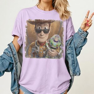 May include: A light purple t-shirt featuring a graphic print of the characters Woody and Buzz Lightyear. Woody is wearing dark sunglasses and a cowboy hat, while Buzz Lightyear stands beside him. The graphic has a distressed, textured border.