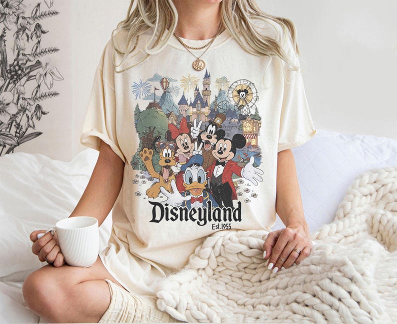 May include: Cream-colored t-shirt featuring a Disneyland graphic with Mickey Mouse, Minnie Mouse, Donald Duck, and Goofy. The graphic includes a castle, Ferris wheel, and fireworks. The text "Disneyland Est. 1955" is printed below the characters.