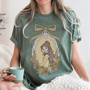 K&ouml;nnte beinhalten: Gr&uuml;nes T-Shirt mit einer Cartoon-Illustration einer Frau in einem gelben Kleid in einem ovalen Rahmen, umgeben von Blumen und einer Schleife. Das T-Shirt hat hochgekrempelte &Auml;rmel und eine lockere Passform.