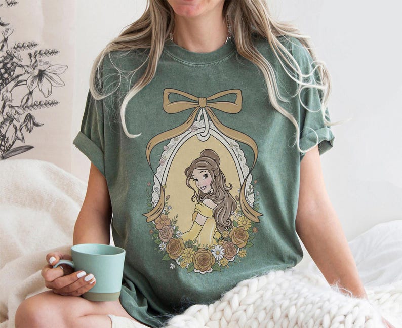 K&ouml;nnte beinhalten: Gr&uuml;nes T-Shirt mit einer Cartoon-Illustration einer Frau in einem gelben Kleid in einem ovalen Rahmen, umgeben von Blumen und einer Schleife. Das T-Shirt hat hochgekrempelte &Auml;rmel und eine lockere Passform.